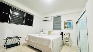 A bedroom with a bed, a bedside table, a wall-mounted air conditioner, a window with blinds, and a framed picture on the wall.