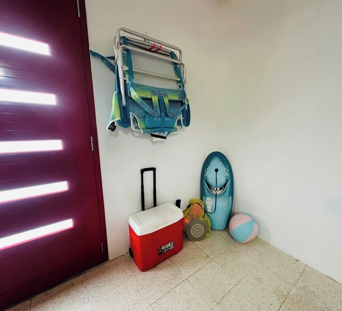 Een kamer met een rode deur, een wandrek met een blauwe en groene handdoek, een rode koelbox, een blauw kussen in de vorm van een haai, en een klein rond kussen.