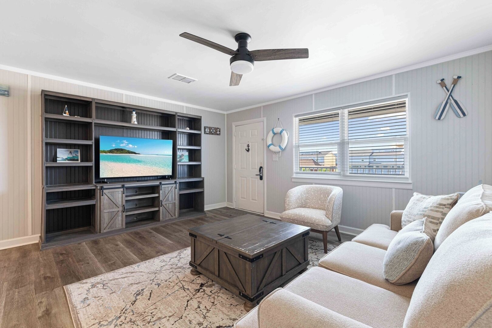A living room with a TV, wooden entertainment unit, ceiling fan, and a large window with blinds.