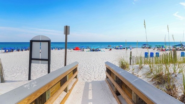 A beach with a wooden boardwalk leading to a gazebo, blue beach chairs, and umbrellas.