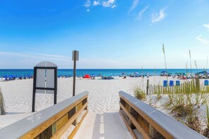 Ein Strand mit einer Holzpromenade, die zu einer Laube führt, blauen Liegestühlen und Sonnenschirmen.