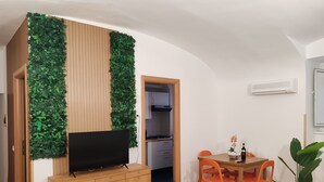 Un espace de vie compact comprenant un meuble en bois, un téléviseur à écran plat, une table à manger avec des chaises orange et un jardin vertical vert.