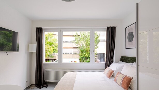 A bedroom with a bed, a window with curtains, a radiator, a lamp, and a framed picture on the wall. - Blueground | Basel-Iselin, view, nr Rhine (Basel)