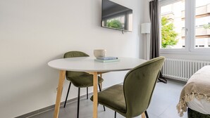 A small, modern room with a round white table, two green chairs, a flat-screen TV mounted on the wall, and a window with a view of buildings and trees. - Blueground | Basel-Iselin, view, nr Rhine (Basel)