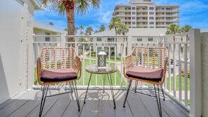 A balcony with two wicker chairs and a small table, overlooking a pool and a building with multiple balconies.