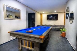 A pool table with balls, a sofa, and a TV in a room with tiled flooring and a mirror.