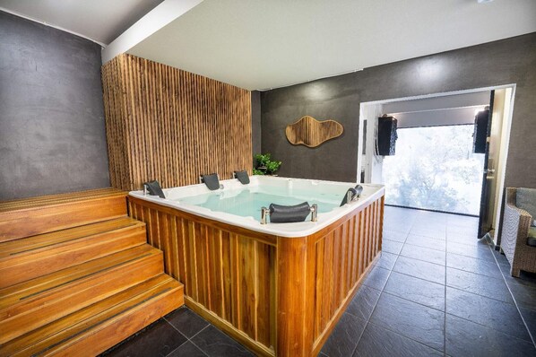 A modern spa room with a wooden sauna, a large freestanding bathtub, and a view of the outdoors through a glass door.