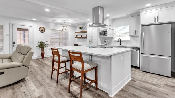 Una cocina moderna con una isla central, muebles de cocina blancos, electrodomésticos de acero inoxidable y un sofá beige en la zona de estar contigua.