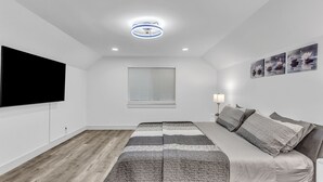 A bedroom with a bed, a flat-screen TV, a ceiling light, a window with blinds, and wall art.
