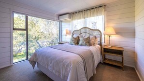 A bedroom with a large window, a bed with a headboard, two bedside tables with lamps, and a view of trees outside. - Villetta Hauraki by Waiheke Pure - Spacious modern home, walk to Palm Beach (Waiheke Island)