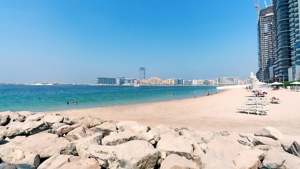Une plage avec un rivage rocheux au premier plan, une étendue de sable et, au loin, la silhouette d’une ville.