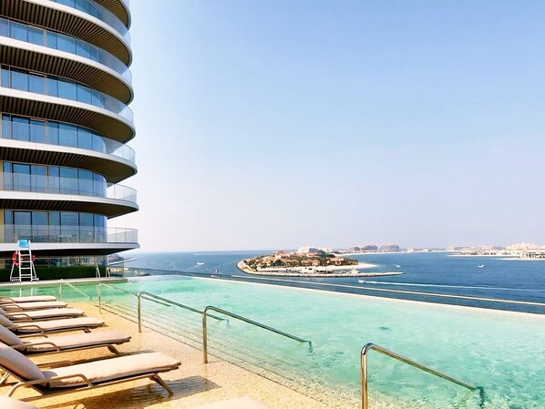 A modern hotel with a curved exterior, a pool area with clear blue water, and a view of the sea and distant buildings.