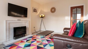 A living room with a flat-screen TV mounted above a fireplace, a colorful geometric rug, a brown leather sofa with cushions, a floor lamp, and a decorative wall clock. - Two bedroom house with free parking, near city centre, sleeps up to five (Edinburgh)