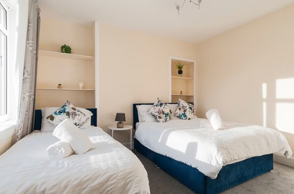 A hotel room with two beds, a small bedside table with a lamp, and a shelf with decorative items.