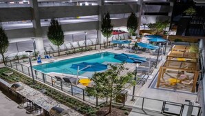 Piscina interior com várias pistas de natação, rodeada por espreguiçadeiras e guarda-sóis, situada dentro de um edifício de estacionamento com vários andares.