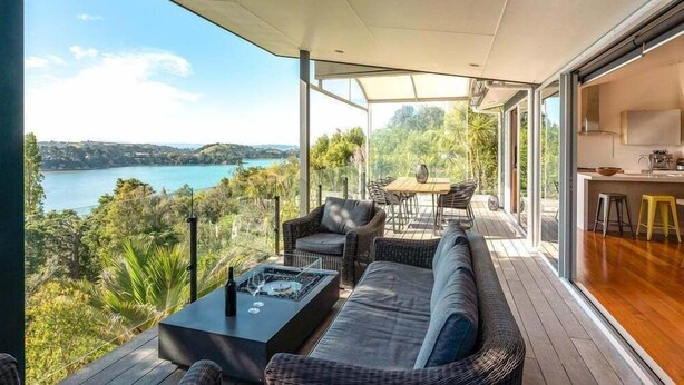 Un espacio al aire libre moderno con sofá, sillas y una mesa de comedor, con vistas a un lago y colinas.