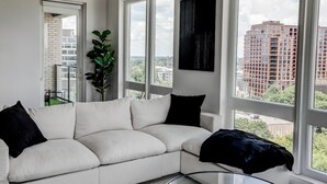 A modern living room with a white sofa, black cushions, a large window with a city view, and a black abstract painting on the wall.
