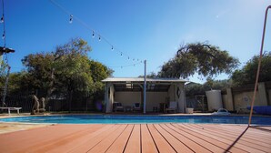 Une terrasse en bois avec une piscine, un espace salon extérieur couvert et une cour clôturée avec des arbres.