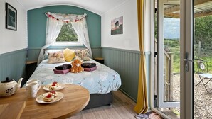 A bedroom with a bed, a window with curtains, a table with plates and a teapot, and a view of the outdoors. - Daisy Shepherd's Hut, North Molton (North Molton)