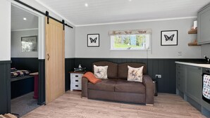 A cozy living room with a brown sofa, a small table, and a window with a view of greenery. - Robins Nest Lodge, South Molton (South Molton)