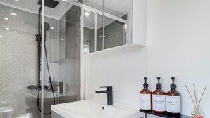 Une salle de bain moderne avec une douche à parois de verre, un lavabo blanc et une étagère sur laquelle sont posés trois flacons de gel douche et un flacon de diffuseur de parfum à bâtonnets.