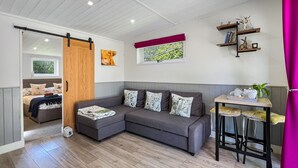 Un salon moderne avec un canapé d’angle gris, une porte coulissante en bois et un petit coin cuisine équipé de tabourets de bar.