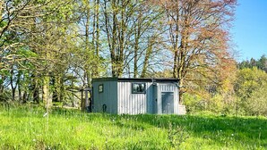 Une petite construction rustique en bois, située dans un champ herbeux, avec des arbres en arrière-plan.