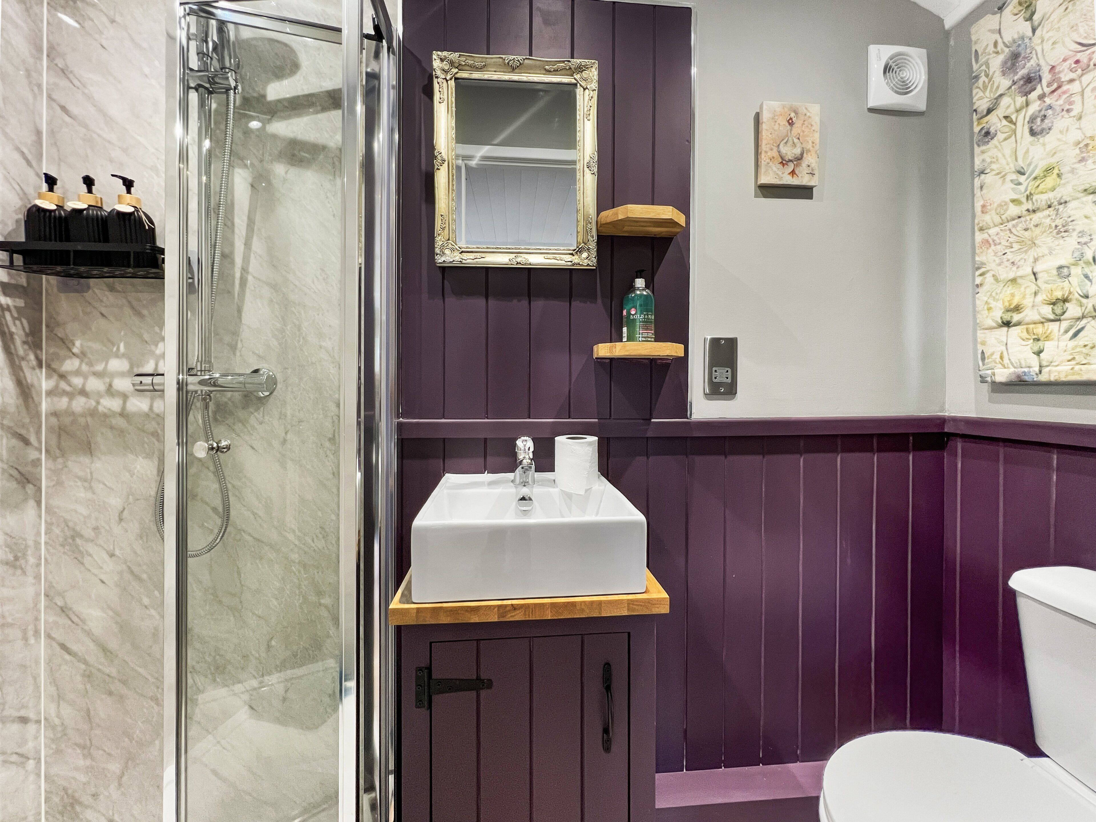 Un baño con cabina de ducha de vidrio, un lavabo blanco con encimera de madera, un tocador morado y un inodoro blanco.