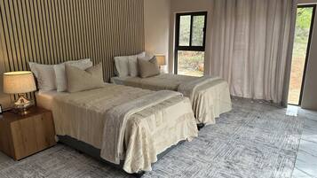 A bedroom with two beds, a nightstand with a lamp, a large window with curtains, and a patterned rug.