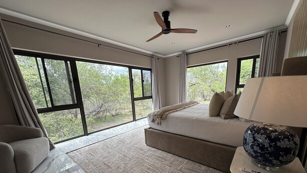 Un'ampia camera da letto con un letto grande, un ventilatore a soffitto e vista sugli alberi attraverso ampie finestre.