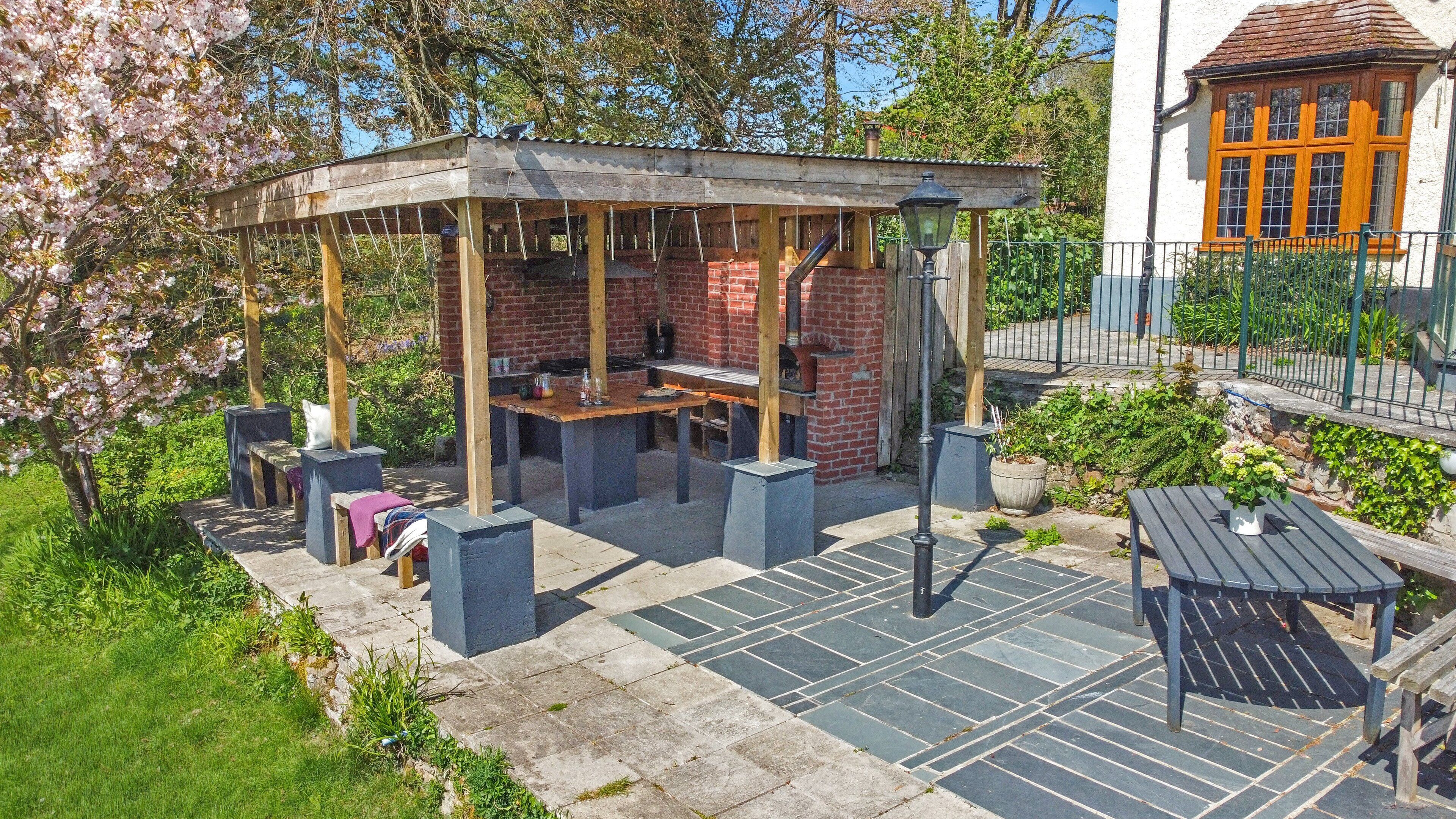 A covered outdoor seating area with a brick barbecue, stone flooring, and surrounding greenery.