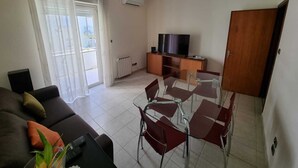 A living room with a sofa, a glass dining table with chairs, a TV on a wooden cabinet, and a sliding glass door leading to a balcony. - COCO Apartments (Catania)