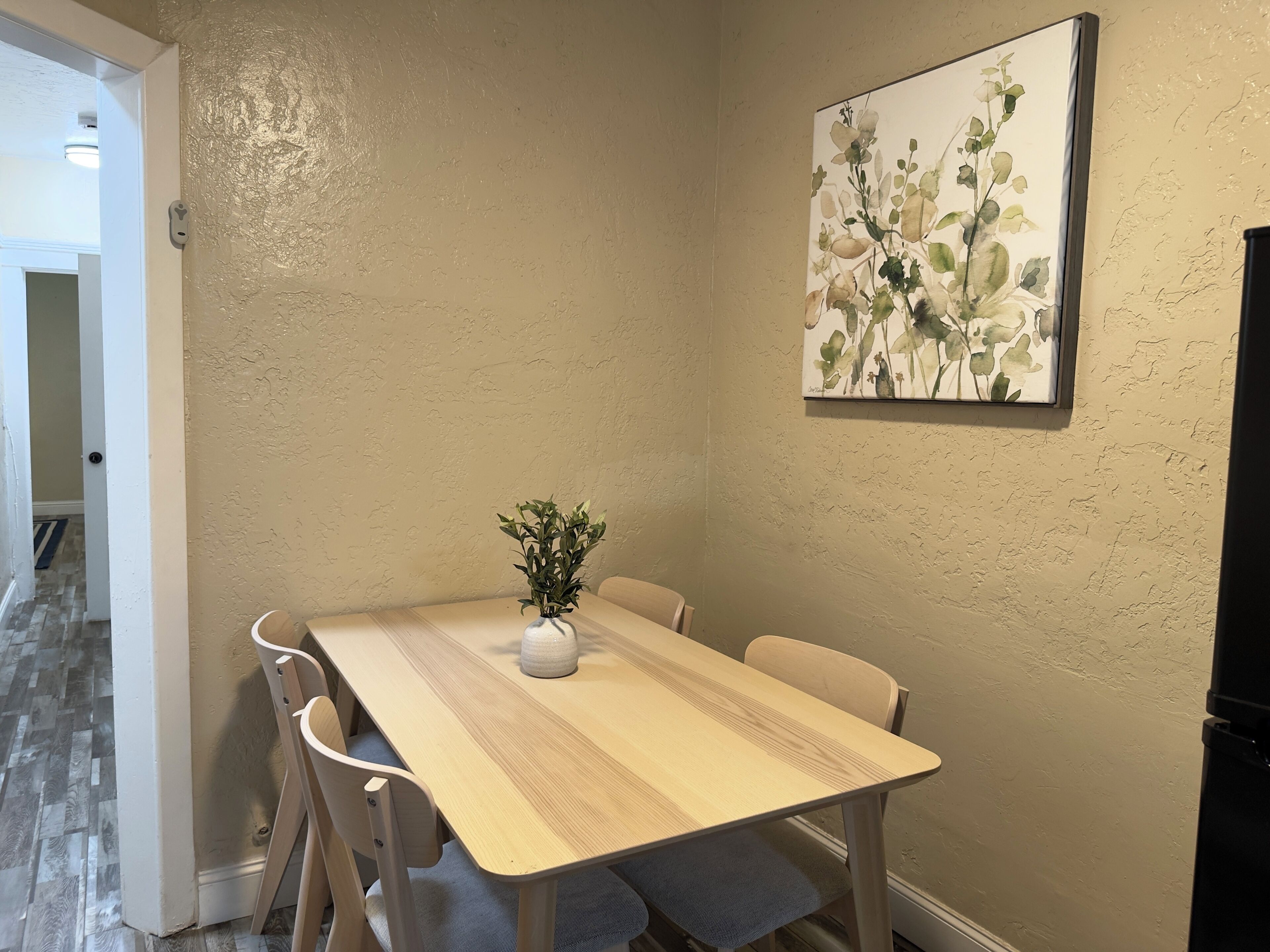 En matplats med ett träbord, fyra stolar, en vas med gröna växter och en blomstermålning på väggen.