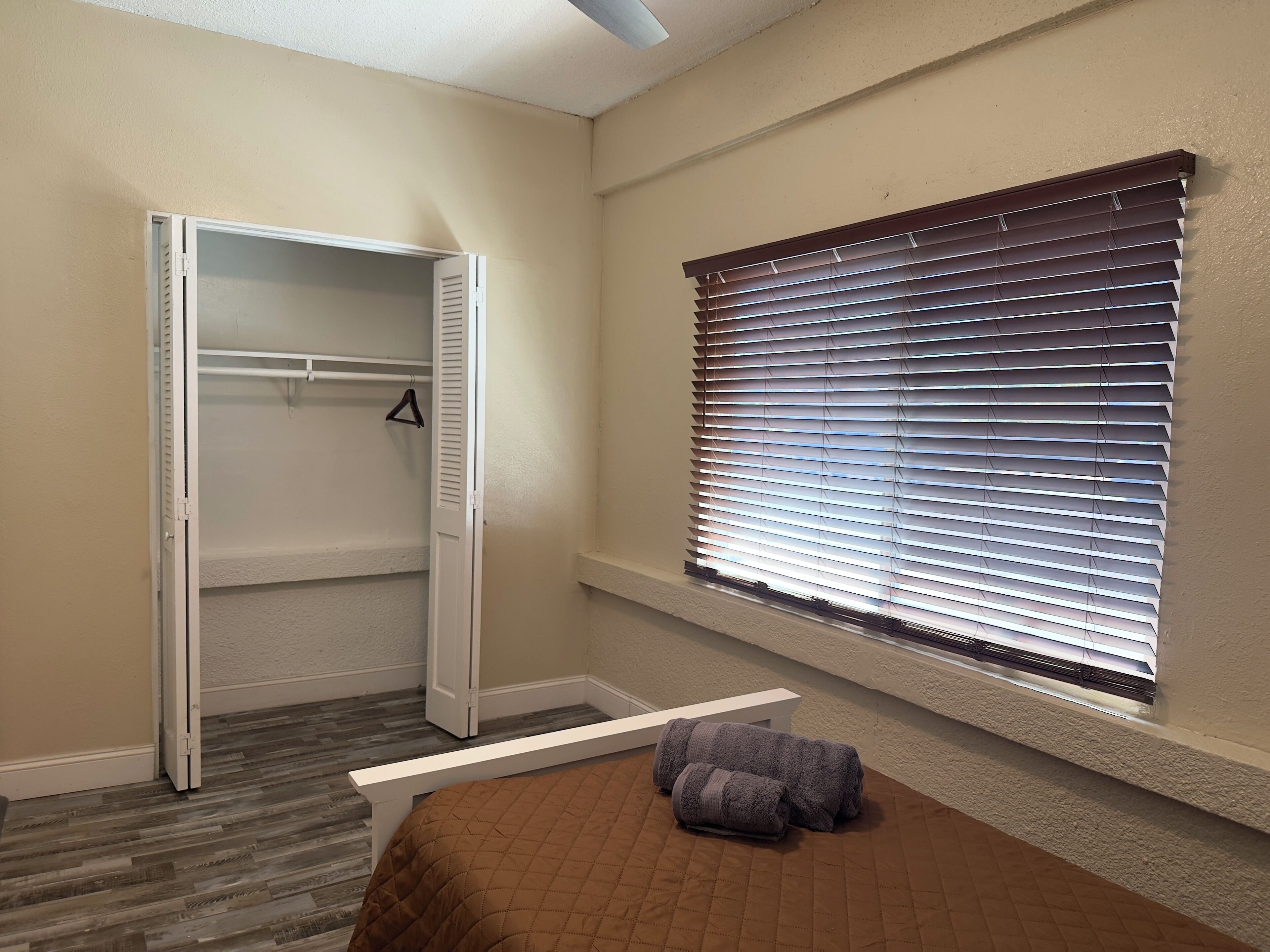 Une chambre avec un placard, une fenêtre avec des stores et un lit recouvert d’une couette marron.