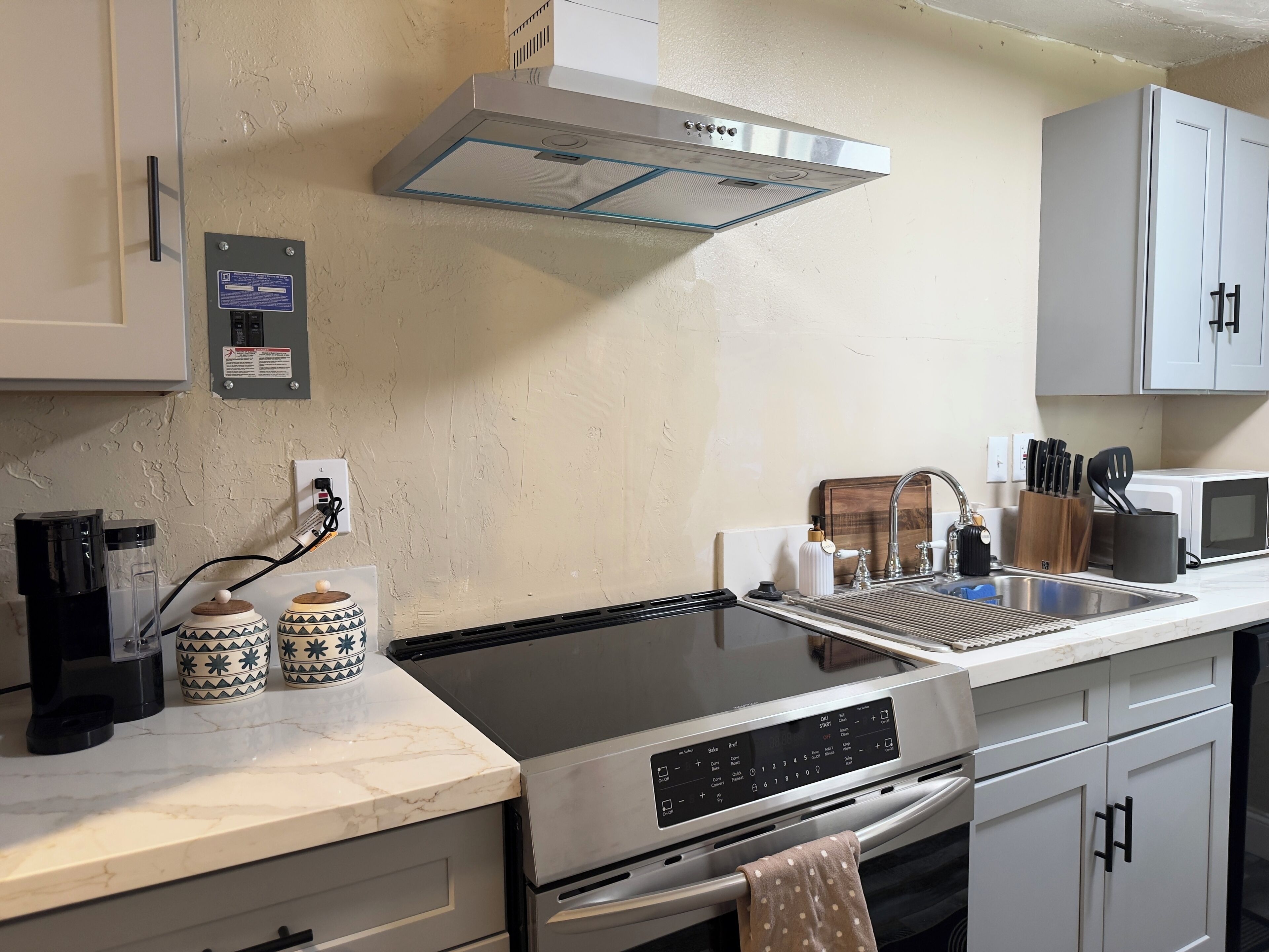 Cozinha equipada com forno embutido, micro-ondas e exaustor em aço inoxidável. Inclui armários, pia e bancada com cafeteira e potes decorativos.