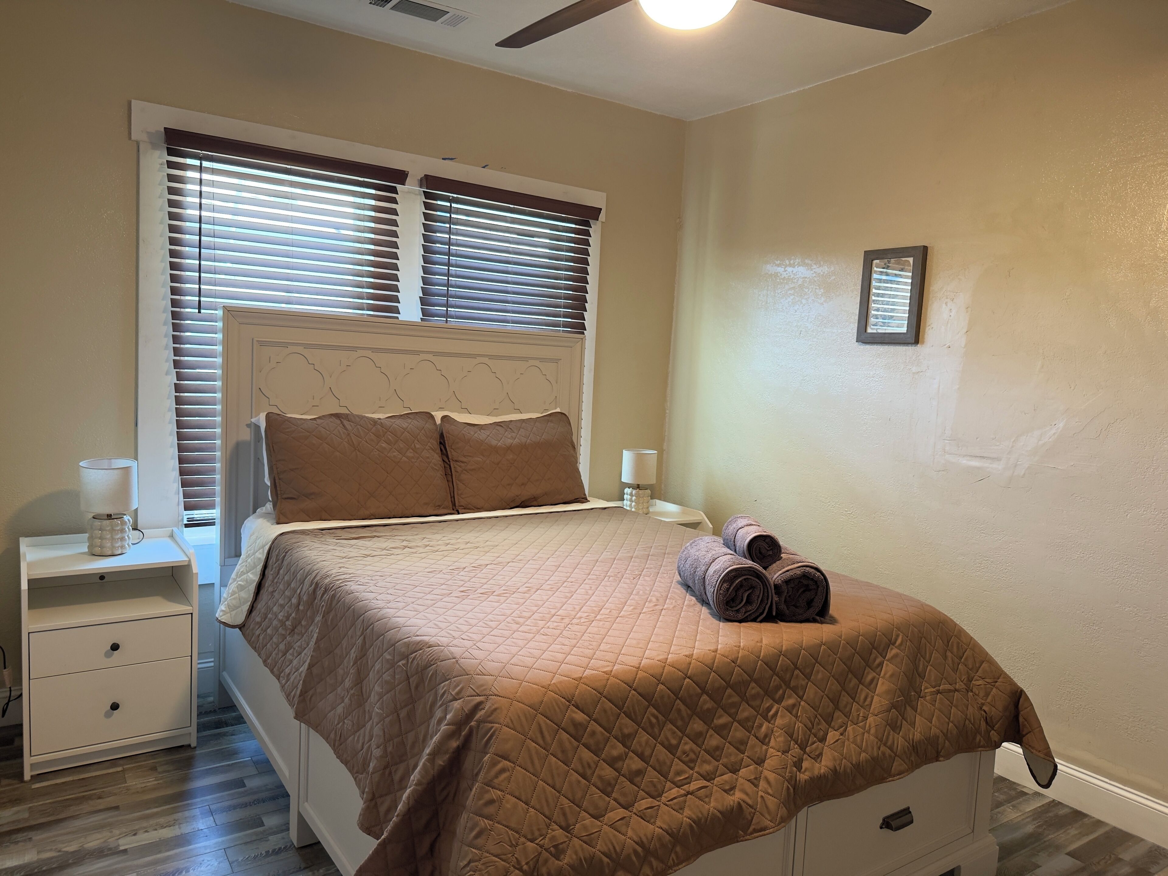 A neatly made bed with a brown quilt, a nightstand with a lamp, and a window with blinds.
