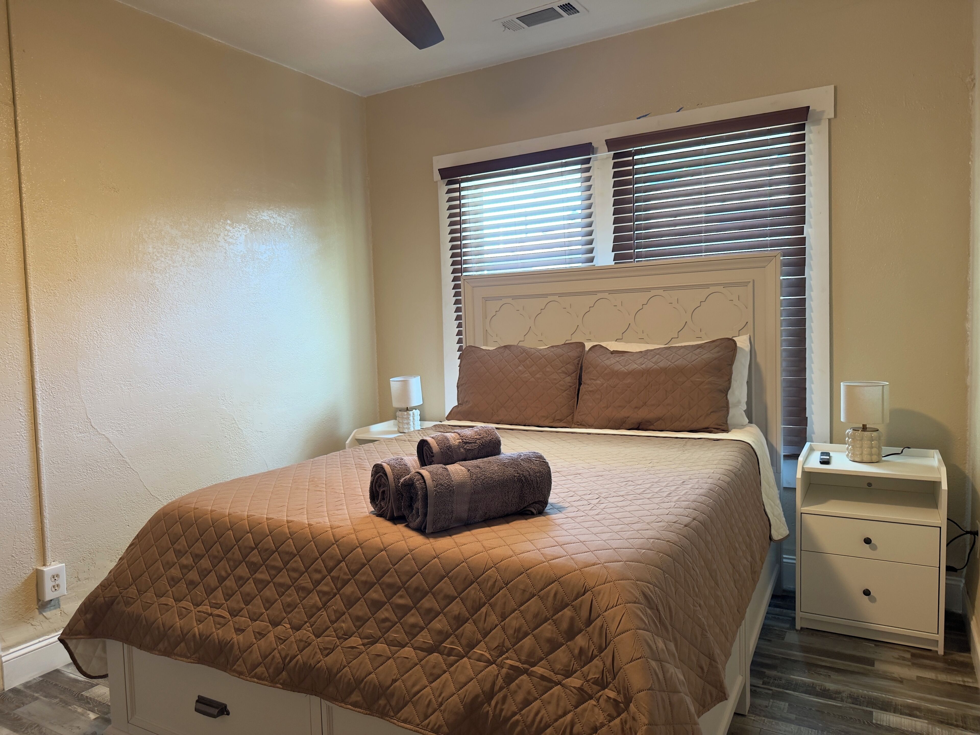 A neatly made bed with a brown quilt, two bedside lamps, and a window with blinds.