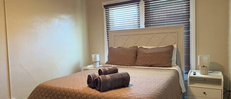 A neatly made bed with a brown quilt, two bedside lamps, and a window with blinds.