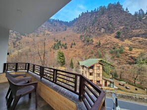 Un balcón con vistas a un paisaje montañoso, un edificio pequeño con techo verde y mobiliario exterior.