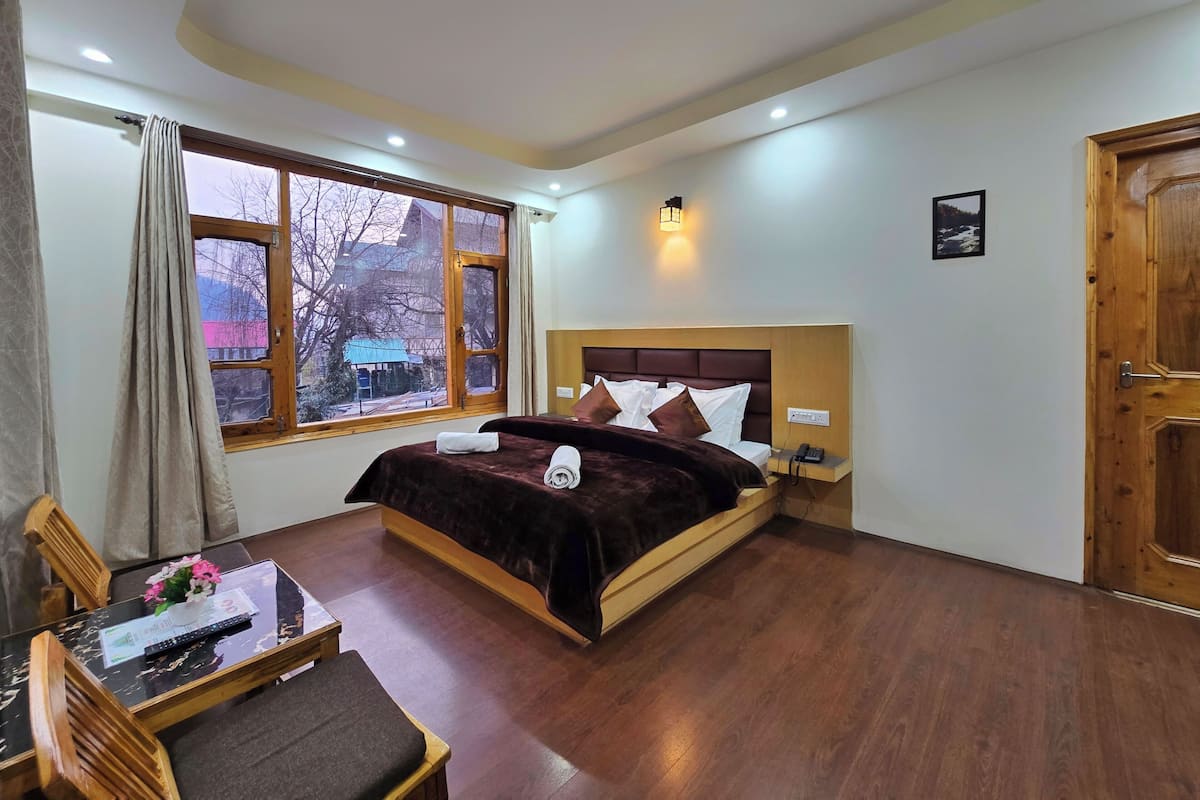 Habitación de hotel con una cama grande, una mesita de noche con un teléfono, una pequeña mesa con un control remoto y flores, y vista al exterior a través de una ventana.
