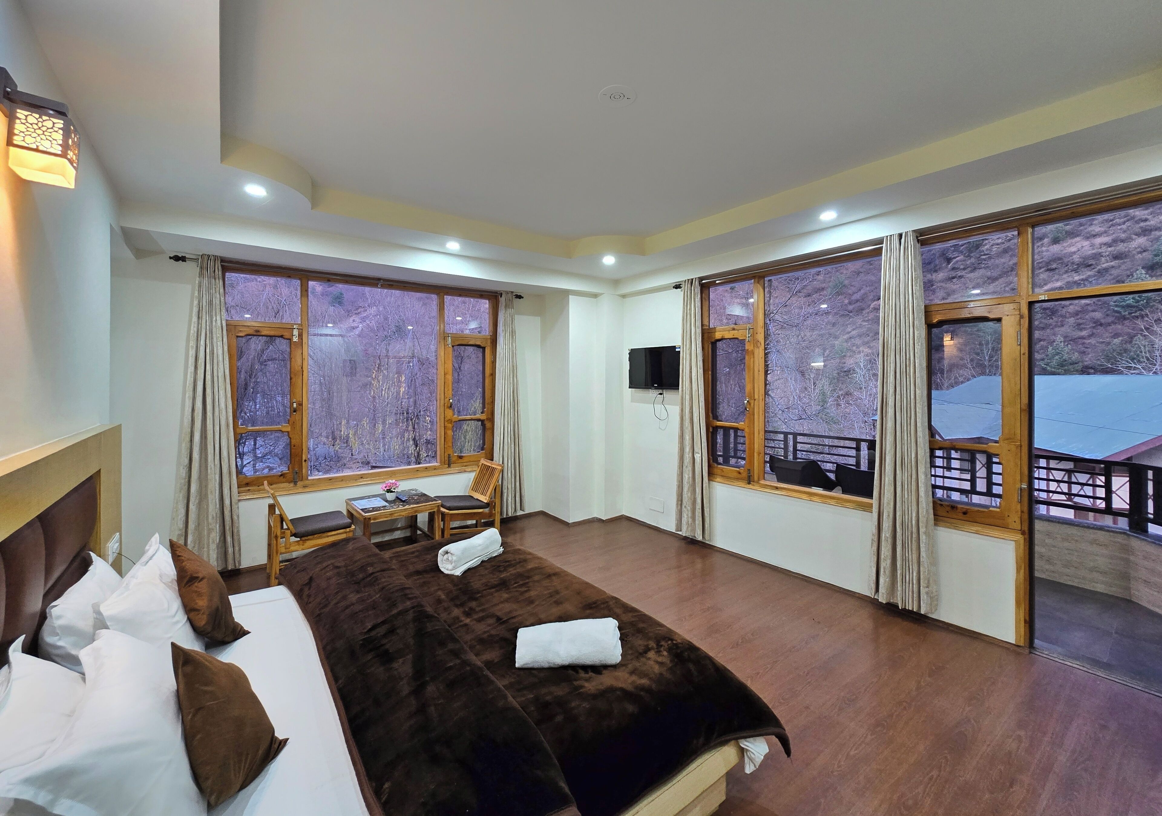 A hotel room with a large bed, a desk, and a TV. There are large windows with curtains, and a balcony with a railing is visible through the windows.