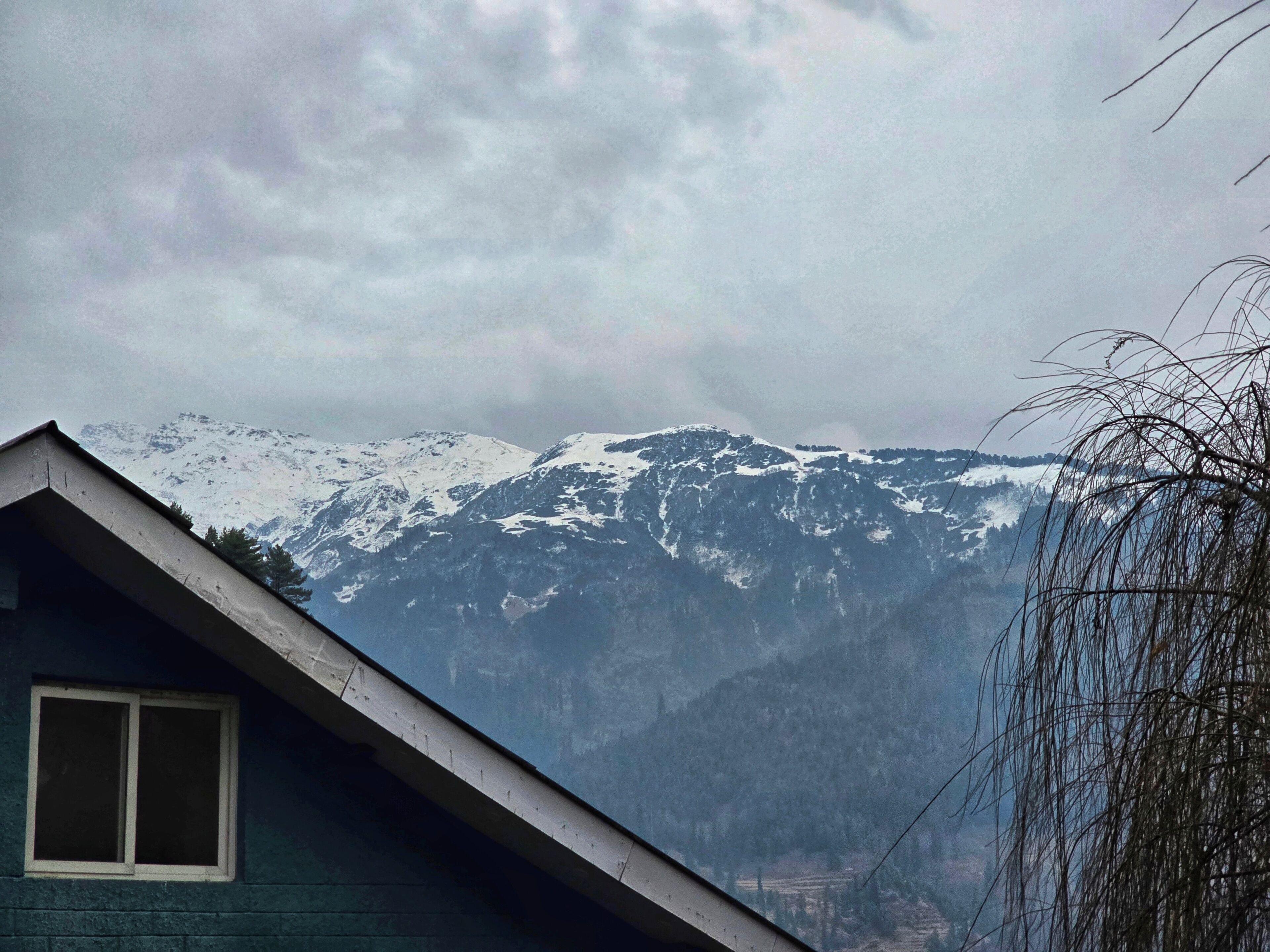 Paisagem montanhosa com picos nevados e uma casa com janela e beiral visíveis.