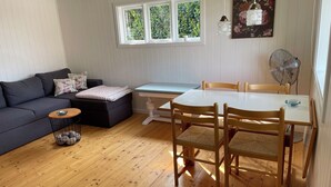 A living room with a sofa, a small table, a dining table with chairs, a fan, and a window with a view of greenery.