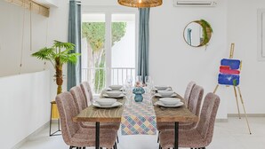 Uma área de refeições com uma mesa de madeira posta para quatro pessoas, rodeada por cadeiras estampadas. Um vaso grande com planta está à esquerda e um quadro em cavaletes à direita.