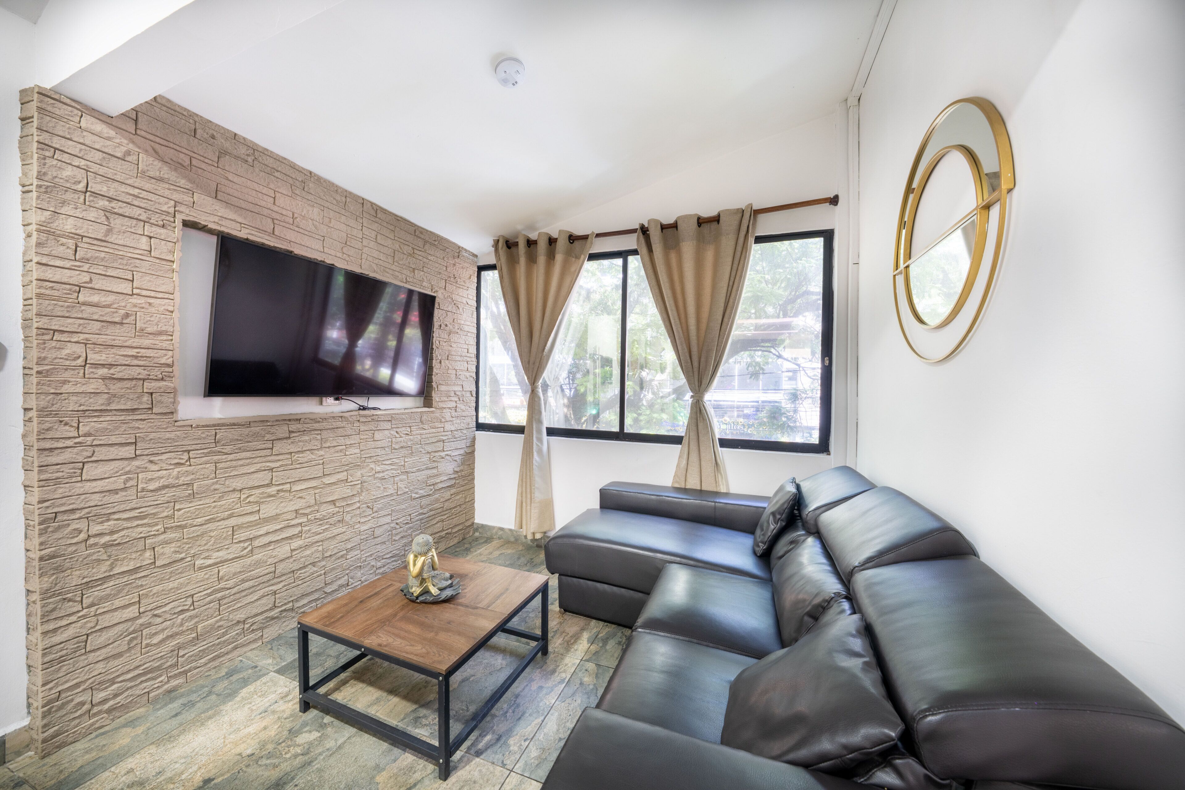 A modern living room with a stone wall, a flat-screen TV, a leather sofa, a coffee table, and a window with curtains.