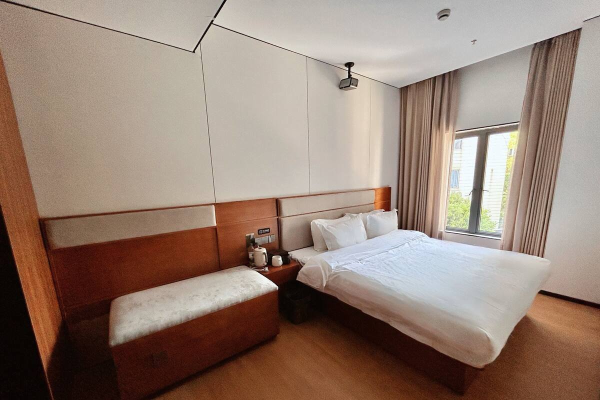 A hotel room with a large bed, a wooden headboard, a bedside table with a lamp, and a window with curtains.