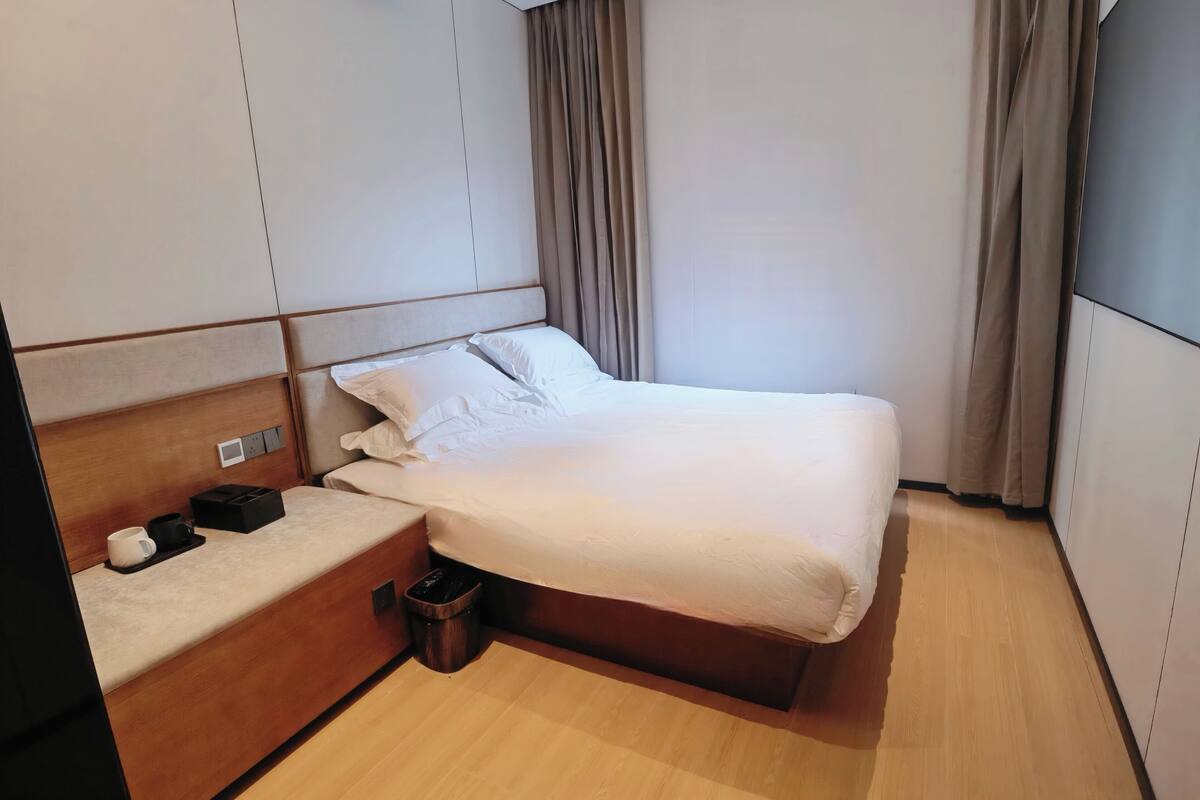 A neatly made bed with white linens, a wooden headboard, and a bedside table with a cup and a black object.