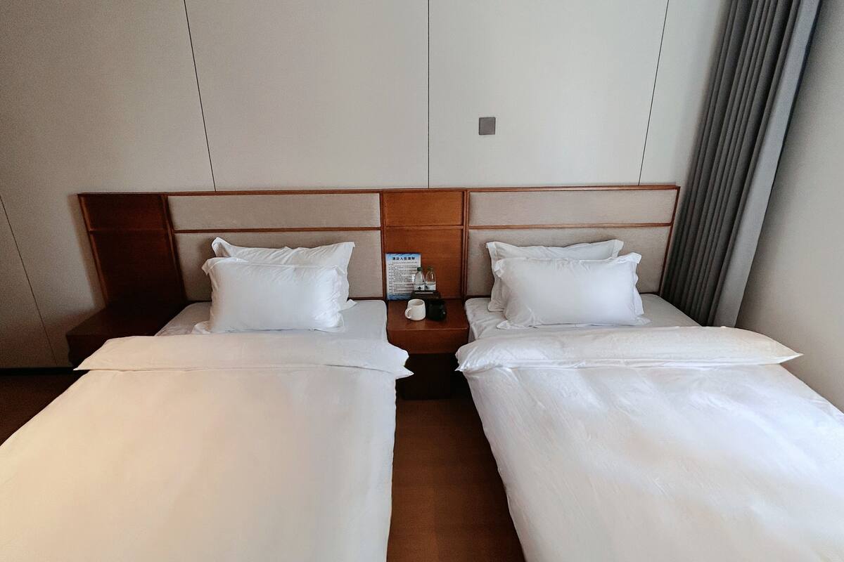 A hotel room with two single beds, wooden headboards, a nightstand with a clock, and a curtain.