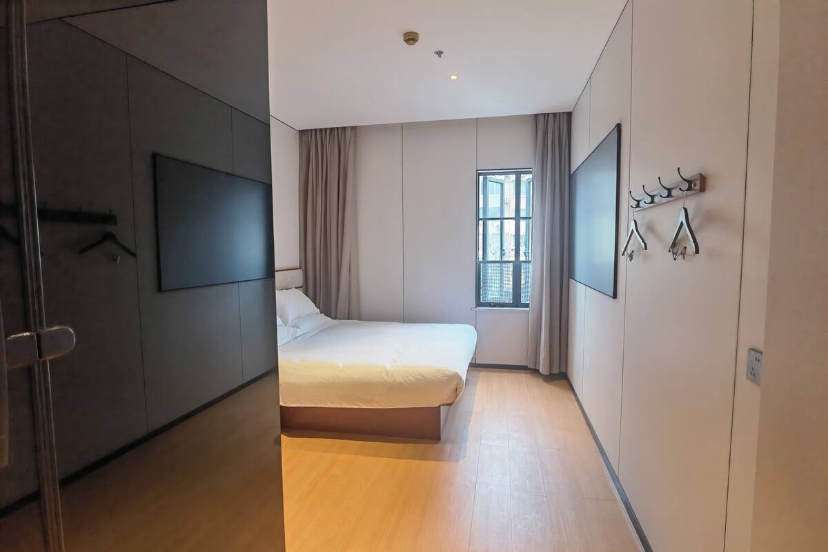 A modern bedroom with a large bed, a TV mounted on the wall, a window with curtains, and a coat rack.