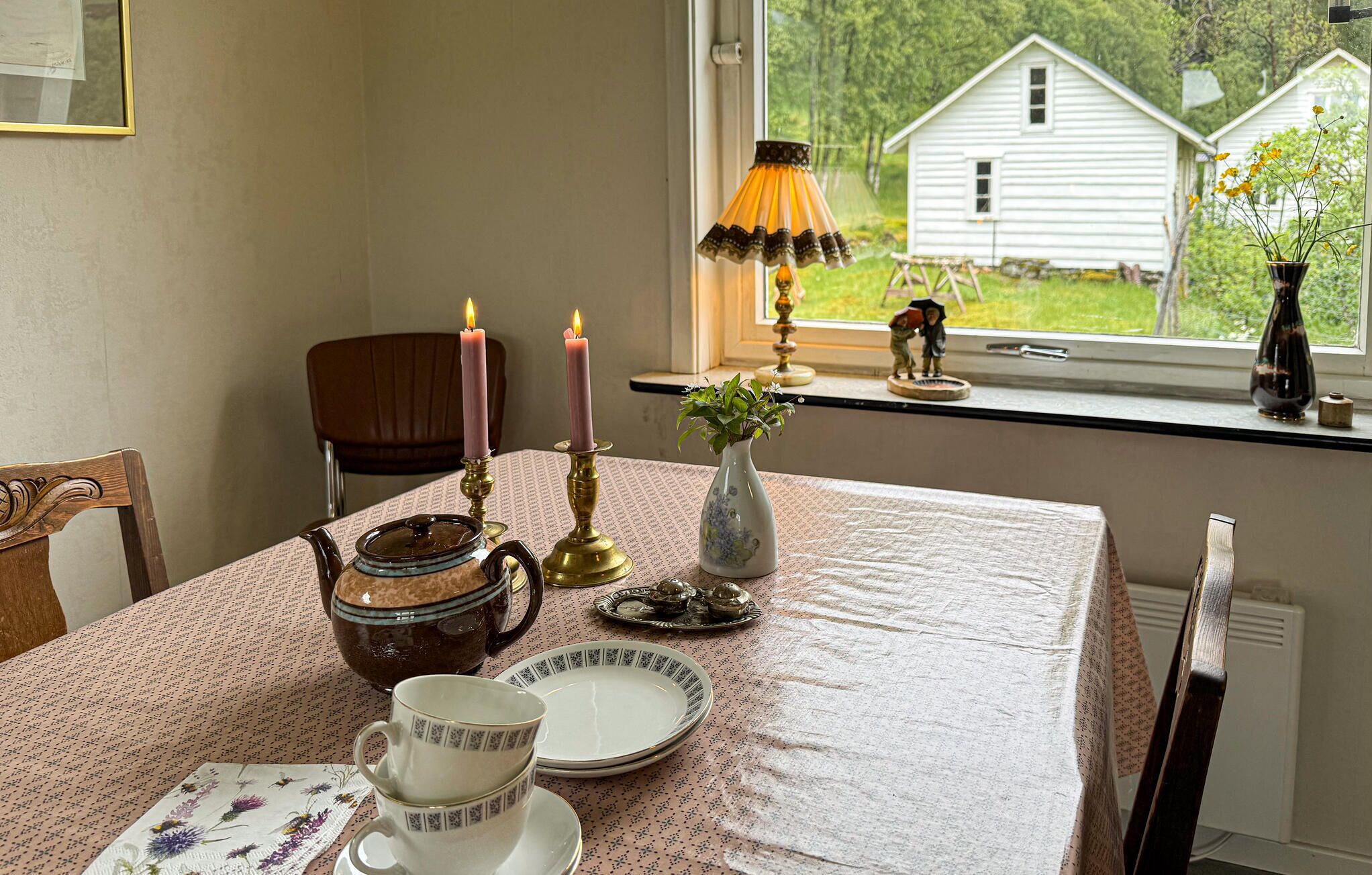 Ett matbord med ett teservis, två ljus, en vas med blommor och en utsikt genom fönstret mot ett vitt hus.